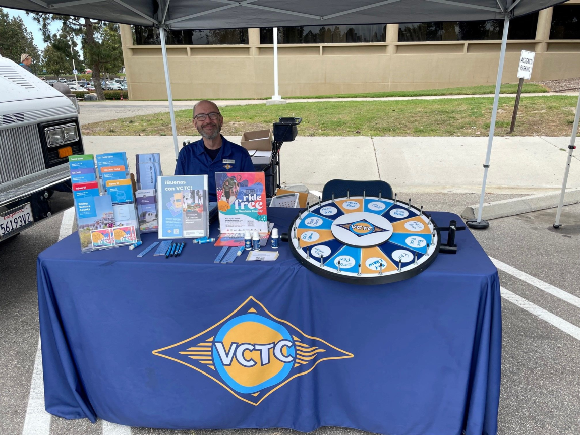 A smiling male volunteer sitting at a VCTC booth with various brochures, swag, and a prize wheel