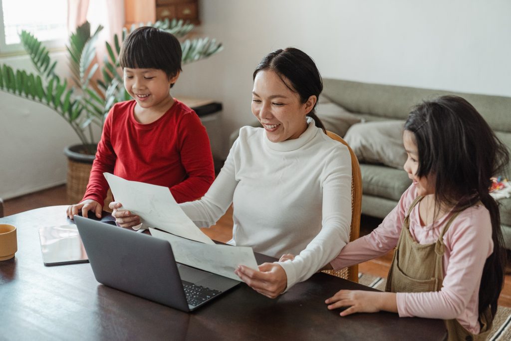 A woman and two children using a laptop and reading papers at home, family activity.