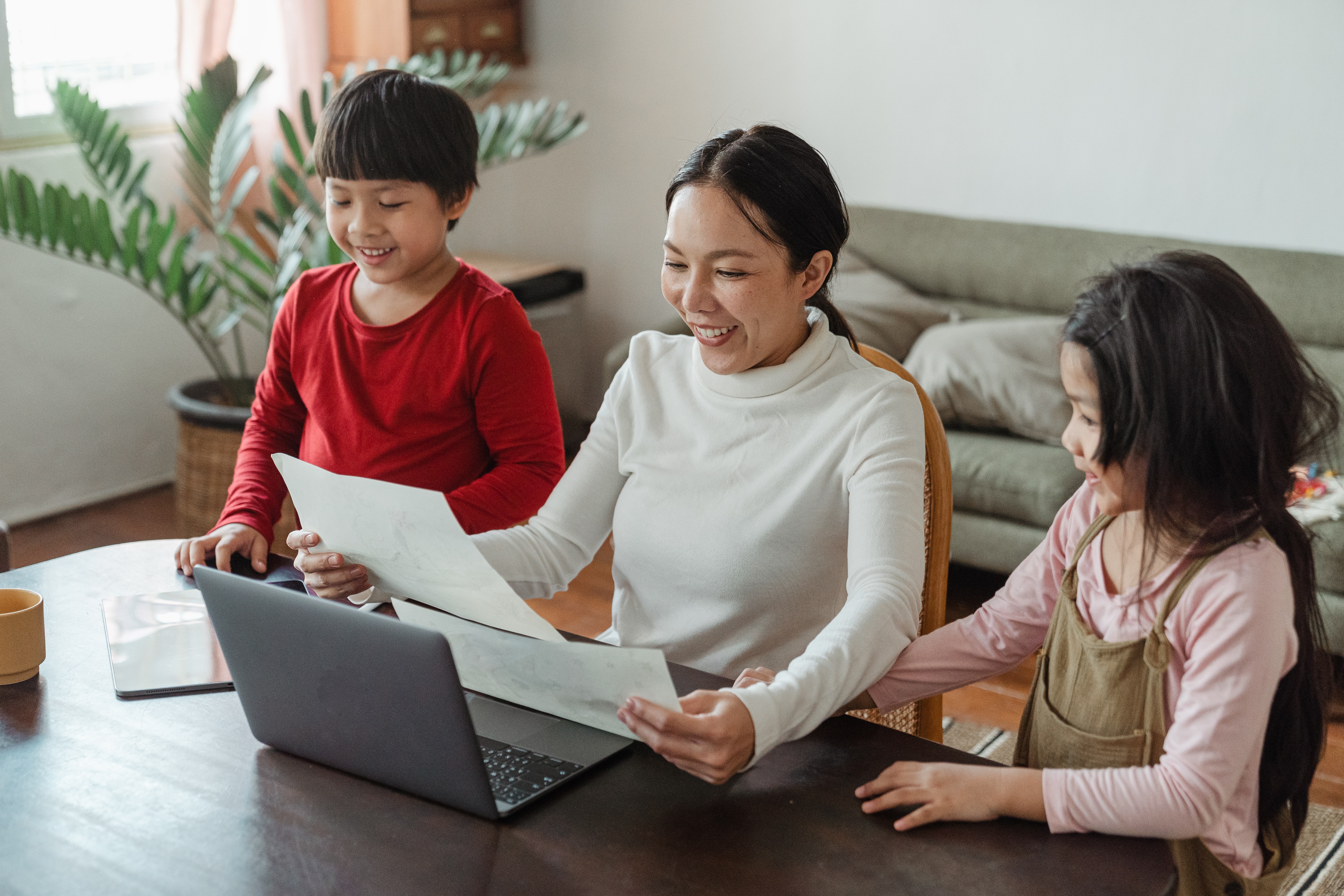 A woman and two children using a laptop and reading papers at home, family activity.