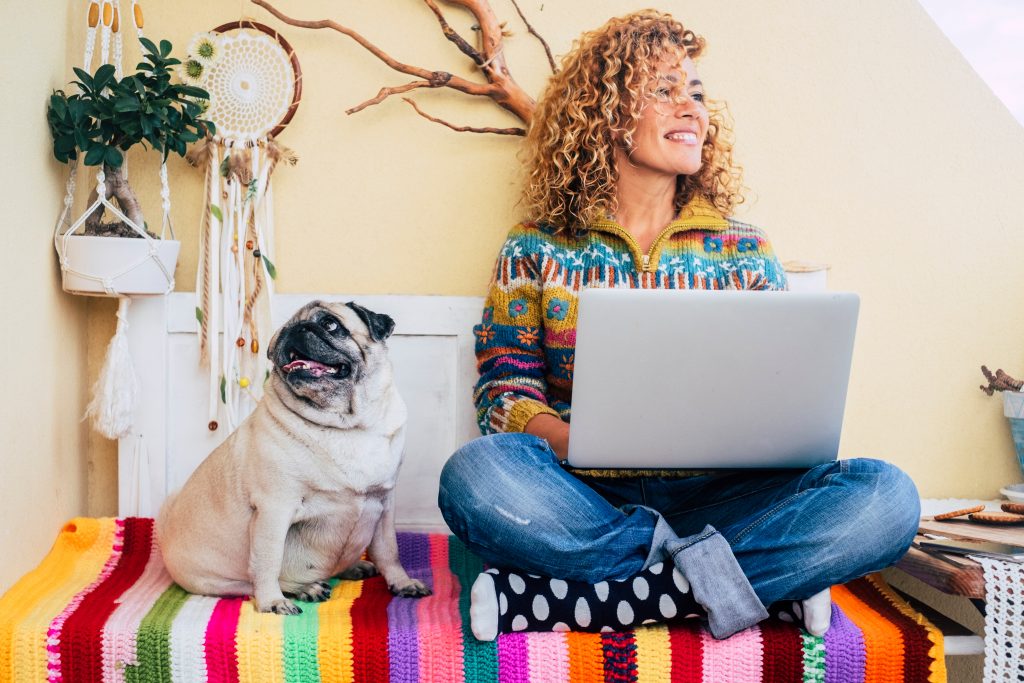 A woman with curly hair working on a laptop with her pug dog indoors, cozy home office setting.