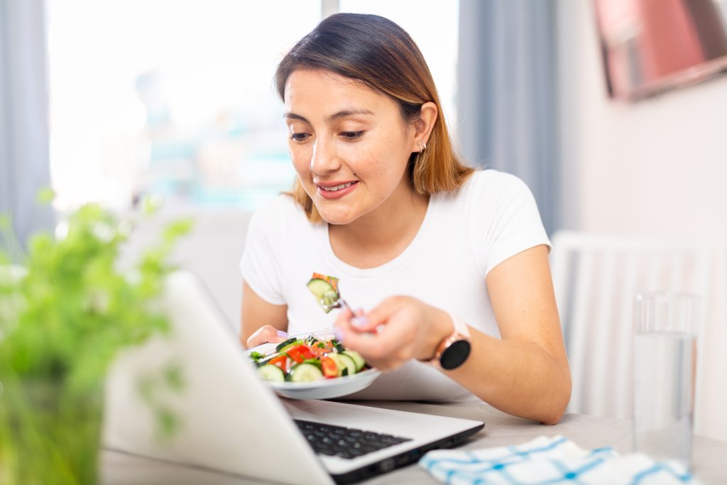 A woman enjoying a healthy salad while working on her laptop at home.