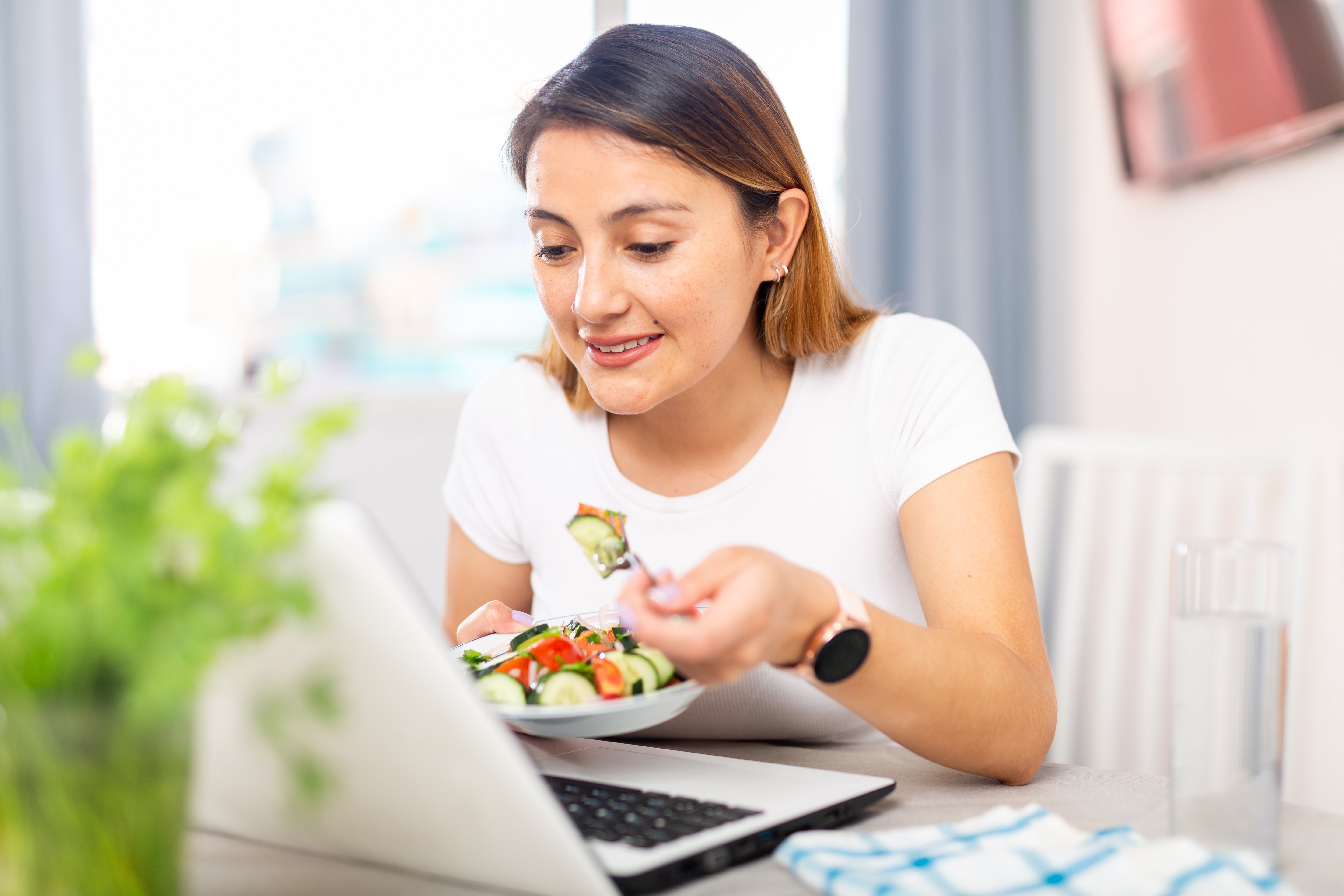 A woman enjoying a healthy salad while working on her laptop at home.