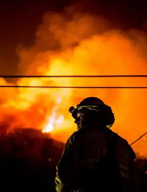 A firefighter confronting a large blaze with thick smoke and intense flames at night.