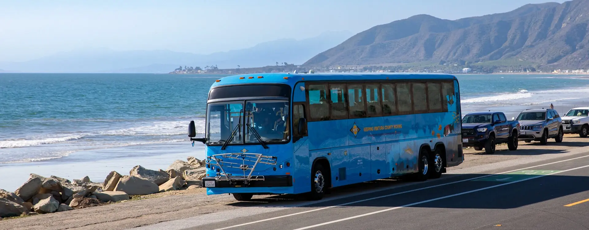 A modern blue bus traveling along a coastal road with mountains in the background.