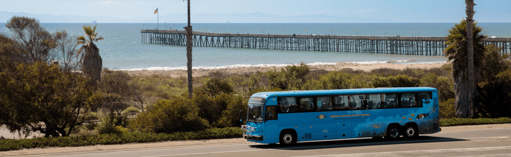 A blue bus traveling along a coastal road with a pier and ocean in the background.