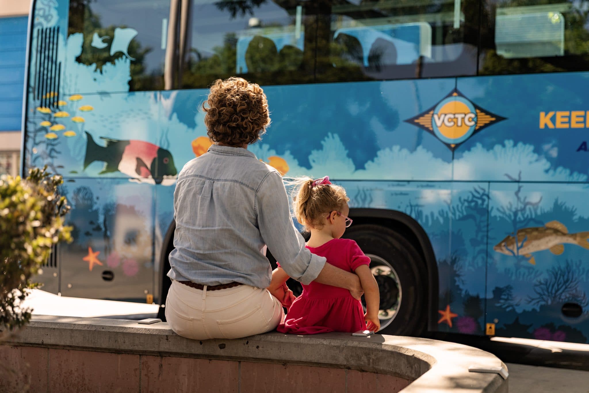Woman and child sitting by a colorful VCTC bus at Goventura Staging.