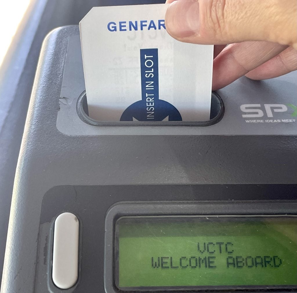A rider inserts a paper transfer slip into the farebox slot on a VCTC Intercity bus. The farebox display reads VCTC Welcome Aboard.
